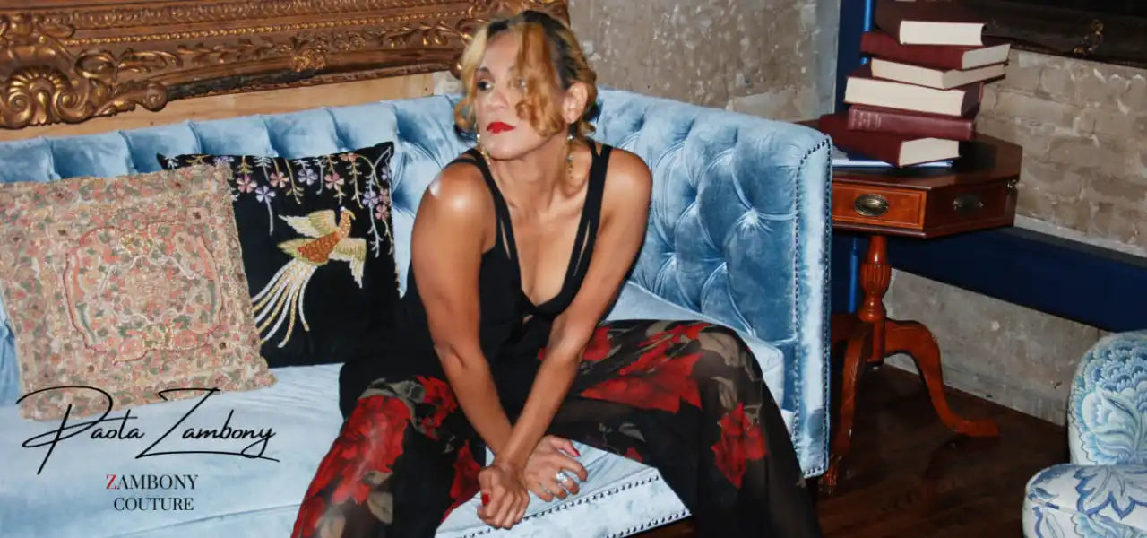 A woman in a black top and floral pants sitting on a blue velvet sofa.