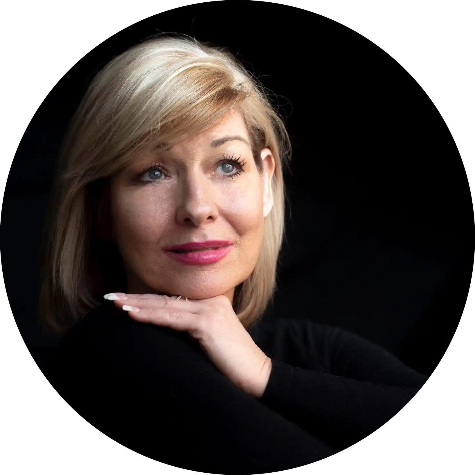 A woman with blonde hair and blue eyes, wearing a black top and pink lipstick, resting her chin on her hand.