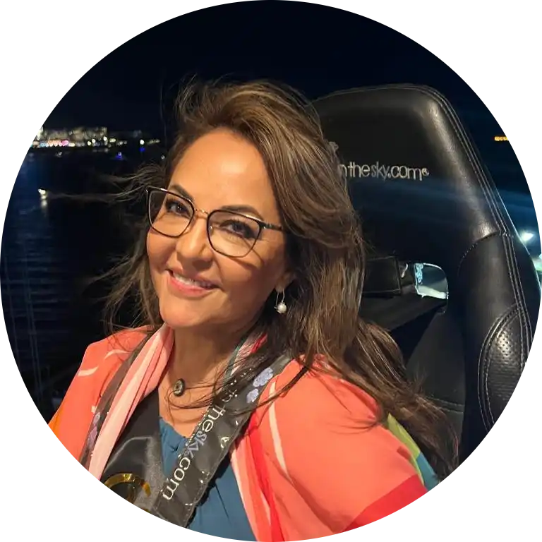A smiling woman with long brown hair and glasses, wearing a peach-colored top and a patterned scarf, sitting in a black chair.