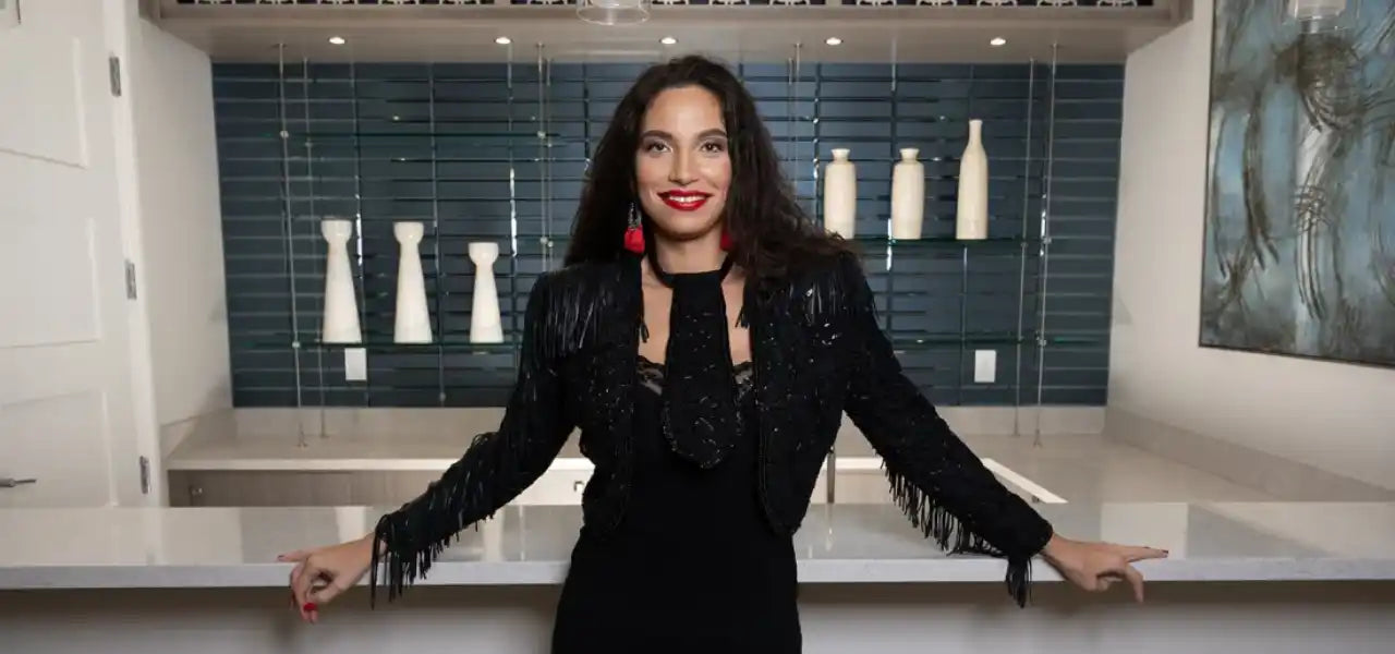 A smiling woman with long dark hair, wearing a black sequined jacket with fringe and a black tie, and red dangle earrings.