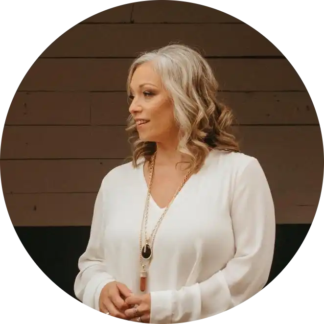 A woman with long, wavy, gray and blonde hair, wearing a white long-sleeved shirt and a long gold necklace with a dark and a light brown pendant.